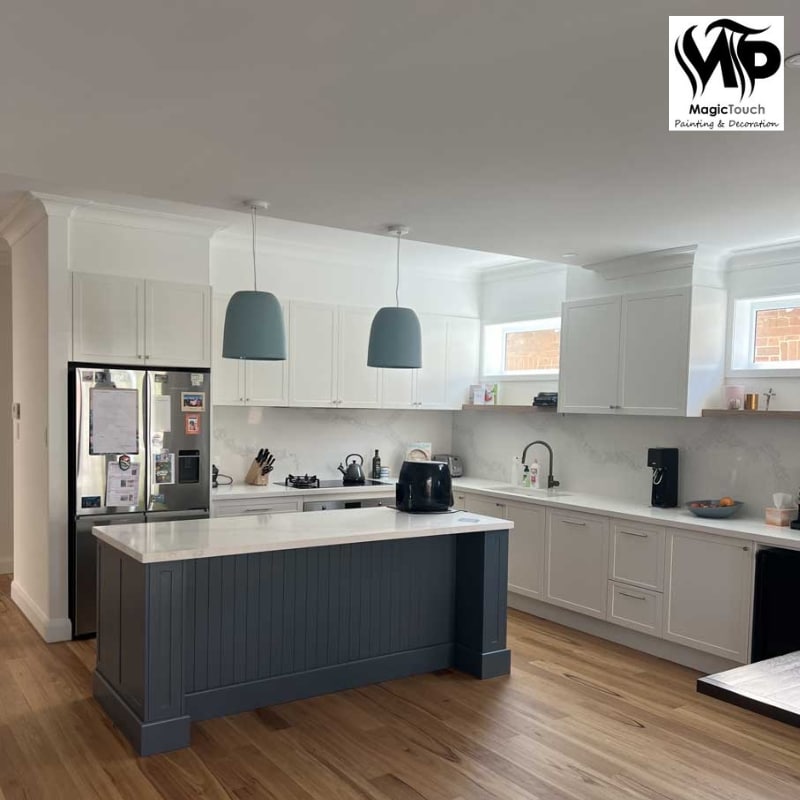 A professional painter in Windsor enhances a modern kitchen with white cabinets, a dark blue island, natural wood flooring, and stylish pendant lighting.